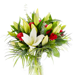 Captured against a clean white backdrop, this arrangement is built around a single, radiant white Oriental lily in full bloom-its thick, creamy petals cupping a pale yellow stamen and a soft green pistil-set slightly forward to draw the eye. Encircling it are numerous unopened and half-open lily buds in lemony green shades that promise unfolding life, while clusters of small, velvety red roses and rosebuds punctuate the scene with dramatic, romantic accents. The bouquet is layered with deep, glossy green foliage that provides weight and silhouette; long, arching palm-like fronds offer motion and a light, tropical whisper as they trail beyond the main mass. Cool-toned eucalyptus stems with their round, silver-green leaves are interlaced for subtle contrast and a refined scent. Everything is gathered in a clear cylindrical glass vase so the pale green stems and fresh water are visible, reinforcing a sense of cleanliness and freshness. The studio lighting emphasises the lilies' satin texture and the roses' plush depth, creating a contrast of softness and structure. From a florist's point of view, this is an arrangement meant to convey heartfelt admiration and ceremonial poise-ideal for anniversaries, congratulations, or a thoughtful sympathy display. Flowers New Barnet often composes similar pieces for same-day delivery across New Barnet and nearby Oakleigh Park or Brunswick Park, where clients ask for the interplay of purity and passionate colour embodied in this bouquet.