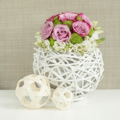 A rounded bouquet of roses and fresh greenery sits serenely at the centre of a white table, capturing the quiet elegance of a New Barnet wedding breakfast just after guests have taken their seats. The flowers form a soft, domed silhouette: large pink and lavender roses, many fully open, reveal layered petals edged with slightly deeper pink tones that give each bloom depth and character. Between them, bright green button chrysanthemums tuck in like little jewels, their compact, textured discs offering a playful contrast to the roses' silky curves. Around the base of the bouquet, clusters of small white filler flowers-each with tiny, star-like petals-create a gentle, ruffled border, suggesting movement as though they are lightly cascading over the rim. The arrangement rests in a striking spherical vase made from white, intertwined rattan-like branches, its open weave casting subtle shadows on the smooth white surface below. To the left, two decorative balls made of pale wicker or wood, one slightly larger, one smaller, echo the same crisscross pattern and bring a crafted, natural feel that would fit beautifully in a home near Victoria Park or along the residential streets around New Barnet station. The background is a softly out-of-focus beige fabric, offering warmth without competing with the flowers' colour. Soft, even lighting washes across the scene, enhancing the fresh greens, romantic pinks, and the intricate textures of the woven materials. The whole composition feels modern yet gentle, an ideal centerpiece for a registry-office reception, anniversary dinner at home, or a romantic bridal table styled by a local New Barnet florist who understands how to keep flowers feeling both refined and inviting.