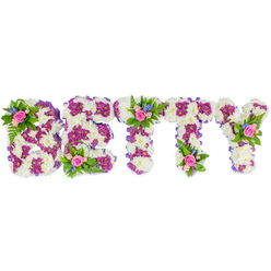 Framed by pure white negative space, this bespoke memorial piece spells BETTY in substantial, block-style letters composed chiefly of creamy white chrysanthemum blooms, their clustered petals forming a plush, tactile surface that suggests quiet comfort. A painterly use of magenta-purple chrysanthemums traces inner shapes and delicate outlines within each letter, creating a thoughtful contrast that reads like a line of memory running through snow. The edges of the letters are softly finished with a narrow purple ribbon, lending a neat, handmade contour. Nestled atop the B, at the centre of the first T and crowning the Y are small posies of soft pink roses-some unfurled, others still cupped-paired with tiny bright blue blooms that echo delphinium or cornflower and lift the palette with a cool, hopeful note. Lush fern fronds and fine green foliage thread through these accents, adding garden-like texture and a fresh, leafy scent in the imagination. The overall design balances tradition and gentle artistry: orderly letterforms softened by tactile blossoms and ribboned edges, ideal for placement at a wake, funeral or celebration of life in the New Barnet community. Imagining it delivered to a chapel near Station Road or placed beside an urn at a local service, the tribute feels both intimate and unmistakably expressive-a floral message of love and farewell for someone known as Betty.