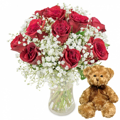 A romantic gift duo is beautifully arranged against a crisp white background, drawing the eye immediately to a full, rounded bouquet of deep red roses displayed in a simple clear glass vase. Each rose is lush and velvety, its petals forming intricate spirals that open outwards in shades of crimson and burgundy, suggesting passion, devotion, and long-lasting affection. Interlaced between the blooms is a generous scattering of white baby's breath, each minute flower like a snowflake resting on a cloud, softening the intensity of the red and adding a light, airy structure. The vase, transparent and unadorned, reveals the neatly trimmed green stems and clear water inside, a subtle reminder of the florist's care and the freshness you might expect from a same-day delivery in New Barnet and the nearby residential streets around Station Road. To the right of the bouquet, a plush teddy bear sits upright, its light brown fur textured and fluffy, almost touchable through the image. The bear's dark button eyes and embroidered features form a sweet, friendly expression, and a dark brown satin ribbon tied in a simple bow around its neck enhances its gift-ready charm. The overall composition feels balanced and intentional, with the strong vertical line of the vase contrasted by the soft, rounded form of the bear. Together, they evoke the feeling of walking into a cosy New Barnet home on an anniversary morning, or opening the door to a surprise Valentine's delivery - a classic pairing for birthdays, apologies, or any moment when words alone are not enough.