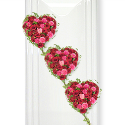In this serene funeral tribute, three rose-filled hearts appear to float in a gentle downward sweep, each one echoing the next in a graceful vertical chain. The hearts are composed of a dense mass of roses in deep crimson red, warm cerise, and soft blush pink, their velvety petals pressed closely together to form a rich, cushion-like surface that feels both luxurious and comforting. Hints of darker shadow nestle in the centres of the fully opened blooms, while the younger, tighter buds introduce subtle peaks and valleys, giving the hearts a sculpted, almost quilted look. Around each heart, slender strands of ivy intertwine and loop, their small, pointed leaves tracing the contours and then spilling downwards to connect to the heart below, creating a living, green framework. The entire display is set against a smooth, bright white background that could easily be the wall of a New Barnet chapel or crematorium chapel, its simplicity allowing the colours of the roses to glow with quiet intensity. There is a gentle, even light across the arrangement, with faint shadows on one side suggesting depth without disturbing the calm, reflective atmosphere. This design feels like a soft-spoken message of love and remembrance, well suited to a farewell service for someone deeply cherished. It would be at home in a New Barnet church near East Barnet Road or placed beside a photo at a memorial gathering, conveying devotion, gratitude, and the promise that memories will remain. The composition speaks of thoughtful care and expert floristry, with every rose and every curve of ivy contributing to a story of connection that endures beyond goodbye.