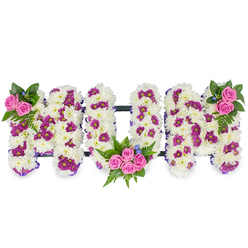 Soft white chrysanthemums form three generous, sculpted letters spelling MUM, each letter built as a three-dimensional cushion of tiny petals that suggest warmth and enduring love. Interspersed through the white mass are pockets of rich purple and magenta spray chrysanthemums that create a subtle dotted pattern-an artisan's technique to add shadow and movement to an otherwise monochrome surface. The arrangement is punctuated by bright pink spray roses used as focal accents: a small, elegant spray sits at the top left of the first M, a more abundant cluster fills the lower curve of the central U, and a mirrored cluster adorns the top right of the second M. Around these roses sit deep green fern fronds and mixed foliage, their texture tactile and their scent faintly green, while tiny blue accent blooms are tucked in like quiet notes of consolation. A discreet dark horizontal bar connects the letters, giving the tribute stability and a finished silhouette ideal for laying near a coffin or placing on a memorial plinth. As a local florist serving New Barnet and nearby streets such as Station Road and Brunswick Park, we craft these personalized tributes with attention to proportion, colour balance and the gentle symbolism families seek at a funeral. This piece reads as both an expression of mourning and a celebration of memory, an arrangement designed to comfort while honouring a beloved mother's place in life and community. Flowers New Barnet prepares each tribute with the sensitivity and care families expect for bereavement occasions.
