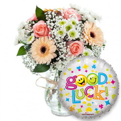 This image captures a comforting 'Good Luck' gift set, described as if offered by a neighbourhood florist on a brisk morning in New Barnet. On the left a clear glass vase, simply tied with rustic twine, supports a full bouquet of peach and pink blooms: silky peachy-orange Gerbera daisies with dark brown centres sit beside pale pink roses whose soft fragrance suggests calm and reassurance. Delicate sprays of white baby's breath weave through the composition like gentle lace, while clusters of small white and lime-green chrysanthemum-like florets introduce a bright, crisp note. Rich dark green foliage provides shadowed contrast and natural depth. In the right foreground a round foil balloon with a metallic silver-and-white sheen cheekily overlaps the vase; scattered hearts, tiny stars and small colourful cross-like accents enliven its surface. The message "Good Luck!" appears in a bouncy, multi-coloured type: the word Good shown in vibrant purple with the two 'o's replaced by yellow smiley faces - one winking, the other sticking its tongue out - and Luck spelled in green, orange, red and blue letters, finished with a green exclamation mark. A discreet KALEIDOSCOPE brand mark sits along the balloon's bottom edge. The arrangement's textures - velvety rose petals, crisp chrysanthemum florets and the glossy balloon skin - combine with a soft, neutral backdrop to create a scene of genuine encouragement, perfect for sending warm wishes across New Barnet toward Brunswick Park, New Barnet Station or nearby homes and offices.