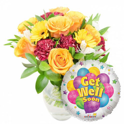 This uplifting get-well composition pairs a lush, hand-arranged bouquet with a shiny round Mylar balloon, styled for maximum cheer and clarity against a white studio background. The bouquet, arranged slightly left of centre in a transparent glass vase so the stems and clear water are visible, is built around large, open roses in warm orange and rich yellow tones whose velvety petals emit a subtle, citrus-honey note. Bright yellow gerbera daisies add bold, sunny faces; deep maroon carnations contribute a textured, almost velveteen contrast; and fine sprays of white alstroemeria create airy punctuation between the stronger colours. A healthy sweep of mixed green foliage - glossy and matte leaves layered for depth - frames and supports the blooms with a natural, freshly-cut look. Nestled to the lower right and overlapping the flowers, the metallic silver balloon features a central cluster of colourful cartoon balloons in sky blue, violet, lime, pink and golden hues, decorated with polka dots, stripes and checks; the message "Get Well Soon" is set prominently in yellow-and-blue lettering with a soft shadow, circled by tiny starbursts and finished with a small "KALEIDOSCOPE" mark at the balloon's edge. The overall impression is warm, optimistic and attentive - ideal for bedside delivery or a doorstep surprise in New Barnet, Brunswick Park or nearby Hadley Wood. Flowers New Barnet prepares arrangements like this with care, ready to brighten a recovery with scent, colour and comfort.