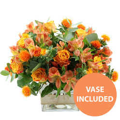A richly coloured floral arrangement in glowing orange and yellow tones is centred against a spotless white background, evoking the warmth of a sunlit afternoon drifting over the streets of New Barnet. The eye is first drawn to the sumptuous orange roses, their velvety petals opening in full, layered spirals, with a subtle wash of yellow emerging towards the heart of some blooms, as if they are gently lit from within. Intermingled with them are sunny yellow garden roses, densely petalled and neatly cupped, adding a sense of abundance and quiet celebration. Around these focal flowers, slender stems of peach and soft orange alstroemeria weave through the design, their delicate, trumpet-like petals patterned with fine speckles and brushstroke stripes, giving the bouquet a graceful, painterly character. Spiky, spherical orange accents-resembling safflower or thistle heads-jut slightly above the softer petals, offering a striking textural contrast that feels playful yet sophisticated. The arrangement is filled out with a rich assortment of greenery: broad, silvery-green eucalyptus leaves fan out in relaxed arcs, while darker green foliage fills the spaces between blooms, creating a lush, layered backdrop reminiscent of mature shrubs in New Barnet gardens near the High Road. All the stems are gathered into a clear, square glass vase, its transparent sides showcasing the clean stems and water line, underscoring the bouquet's freshness. A wrap of natural beige burlap or jute encircles the vase's middle, lending a rustic, homely charm well suited to a local New Barnet home. In the bottom right corner of the image, a vivid orange circle carries the words "VASE INCLUDED" in bold white capitals, making it clear that this warm, cheerful bouquet is a complete, ready-to-display floral gift, perfect for birthdays, housewarmings, or a heartfelt thank you delivered locally.