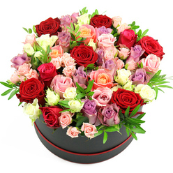 A chic black hat box with a subtle red trim sits isolated against a pure white background, overflowing with an opulent, dome-shaped bouquet of roses that looks as if it has just been arranged on a New Barnet florist's workbench. The round box, with its deep, velvety finish, offers a dramatic contrast to the mass of colour above it, much like the juxtaposition of busy New Barnet streets and the calm greenery of Hadley Common nearby. The roses are packed closely together in a carefully curated tapestry of shades: dark crimson blooms hint at intense romance, their petals lush and velvety, while rich fuchsia and deep pink roses add a lively, celebratory energy. Interspersed among them are softer tones of blush and nude pink, gentle peach and coral, and a scattering of creamy whites that catch the light and keep the composition bright and fresh. Cool lavender and muted lilac roses thread through the warmer colours, adding finesse and a slightly whimsical feel. Some roses are fully open, revealing intricate, layered centres, while others remain in tighter buds or small spray clusters, giving a natural rhythm and depth to the dome. Slim stems of greenery, possibly eucalyptus or fern-like foliage, peek through the petals, offering a crisp green accent that keeps the palette from becoming too heavy. The lighting is bright and even, highlighting the subtle differences in petal texture-from satiny smooth to slightly ruffled edges. This arrangement radiates luxury and care, ideal for a Valentine's surprise in New Barnet, a special birthday delivery near East Barnet Village, or any occasion where you want the flowers themselves to do the talking with grace and abundance.