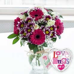 A lush, celebratory floral gift arrangement rests on a clean, pale surface, as if waiting to be handed over at a New Barnet doorstep. At its heart, bold magenta gerbera daisies fan out their wide petals around dark central disks, creating an eye-catching focal point that feels both modern and joyful. Around these key blooms, smaller chrysanthemums display intricate rings of deep purple that fade to snow-white tips, each flower finished with a bright yellow centre, like tiny bursts of sunlight. Cream-toned white roses nestle carefully among the gerberas and chrysanthemums, their layered petals still slightly closed, suggesting freshly cut garden blooms arranged just this morning. Fine sprays of white baby's breath weave between the larger flowers, softening the edges and lending the whole bouquet a delicate, cloud-like halo. Rich green leaves, some glossy and rounded, others more slender, add structure and depth so the design appears full and rounded from every angle. The flowers are gathered into a clear glass vase filled with water, the stems trimmed evenly and visible through the glass, giving a sense of natural simplicity. A piece of rustic twine is tied neatly around the vase's neck, from which hangs a small white tag printed with a pink rose symbol and green handwriting-style text that hints at hand-picked care. To the right, a reflective silver heart-shaped foil balloon catches the light, its white centre printed with playful multicoloured letters that spell out a loving message offered "24-7." The softly blurred, bright background, reminiscent of a sunny window above a New Barnet garden, gives the image a gentle, uplifting feel, ideal for conveying affection, encouragement, or celebration through local flower delivery.