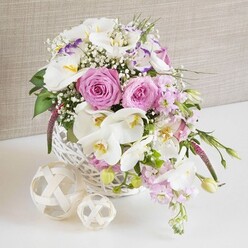 A round white wicker basket, placed on a clean white surface, holds a lush, carefully balanced floral arrangement that feels perfect for a relaxed but elegant wedding breakfast in New Barnet. The basket itself is rustic yet refined, its tightly woven strands forming a soft, circular cradle for the flowers. Inside, pastel pink roses open generously, their layered petals curling into classic spirals that speak of romance and long-planned celebrations. Between them glide white Phalaenopsis orchid blooms, their smooth, wing-like petals and warm yellow centres adding a touch of luxury that would not look out of place in a boutique venue off Station Road. Ruffled white lisianthus, some kissed with a fine edge of lavender-purple, weave in and out of the larger flowers, while stems of pale pink stock arc gently towards the table, suggesting an easy, natural movement as if the flowers had just been gathered from a local New Barnet garden. Wisps of baby's breath scatter through the design like tiny stars, creating a soft haze that lightens the whole piece. Fresh green foliage peeps through, with a few slender leaves and buds reaching beyond the basket's rim to keep the arrangement from feeling too formal. To one side rest two small decorative spheres made from light, woven reeds, echoing the basket's texture and adding to the organic, handmade mood. Behind everything, a neutral, lightly textured beige background provides a calm setting, allowing the soft pinks and crisp whites to glow. The scene suggests an intimate celebration, a thoughtful wedding table arrangement, or a refined gift for same-day delivery to homes and venues around New Barnet.