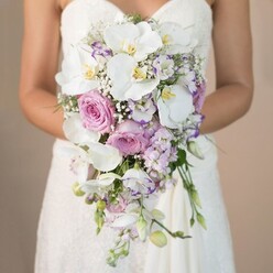 The photograph reveals an elegant bride in New Barnet, her presence bathed in the gentle glow of ambient light streaming in like the first warmth of a spring day at Oak Hill Park. She holds at her waist a splendidly crafted, cascading bouquet that draws the eye with its serene combination of white, blush, and soft purple shades. At the bouquet's core, expansive white phalaenopsis orchids with golden hearts radiate understated glamour, their satiny petals fanning outward with remarkable composure. Soft pink garden roses, full and lush as if freshly picked from a nearby New Barnet garden, nestle among the orchids, their romantic aroma evoking memories of bridal mornings. Interspersed throughout, slender stocks of lavender and tendrils of ruffled eustoma-some edged ever so slightly in deeper purple-introduce a tactile tapestry of spiky and curled textures. Billowing clouds of white Gypsophila add a whispery volume that allows the eye to drift through the bouquet's gentle cascade, reminiscent of light breezes along East Barnet Road. Minimal but deliberate foliage peeks through, grounding the bouquet in natural freshness. The bride's simple yet ornate strapless gown, with its lacework shimmering subtly, frames the flowers and draws attention to the artistic skill behind this arrangement. Every petal and frond seems imbued with the anticipation and joy of New Barnet wedding mornings, brought to life by the dedication of local florists for couples seeking something truly exquisite for their special day.