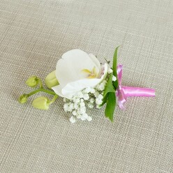 A single white Phalaenopsis orchid boutonniere rests horizontally on a pale oatmeal-coloured, linen-textured cloth, the neutral backdrop allowing every floral detail to stand out with quiet confidence. The orchid bloom is pristine and luminous, its broad, rounded petals forming an elegant open cup, velvety and almost porcelain-like in the way they catch the soft, even light. At its centre, touches of soft yellow and muted pink create a gentle glow, adding warmth and a sense of intimacy reminiscent of a calm morning in New Barnet before guests arrive for a ceremony. Around the orchid's base, wisps of white baby's breath gather like a fine mist, their tiny clustered blossoms creating a light, clouded texture that softens the edges of the design. Slender, fresh green leaves emerge discreetly from behind, their smooth, tapered shapes and vivid colour bringing a natural, garden-fresh contrast to the white blooms. Extending to one side, a delicate stem bears a pair of tightly closed, lime-green orchid buds, each bud promising future blossoms, echoing the hope and new beginnings celebrated in weddings and proms across the local area. The stems are carefully wrapped in a sleek band of bright fuchsia pink satin ribbon, tied into a neat bow whose glossy finish introduces a joyful, modern accent. The light picks up the faint sheen on the ribbon and the subtle textures of fabric and petal, creating an overall feeling of poised, romantic refinement-perfect for pinning to a lapel at a New Barnet wedding, civil ceremony, or any elegant evening event.