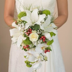 The image captures a New Barnet bride holding an extraordinary cascading wedding bouquet, its beauty revealed in exquisite detail. At its core, several generous white lilies, their petals marked by gentle cinnamon freckles, exude an air of bridal purity and charm. Resting within is a single cream-hued rose, its delicate, tightly gathered petals a timeless symbol of romance. The bouquet's distinctive flow is crafted by strands of white Phalaenopsis orchids, each floral cascade adding a sense of movement, much like the gentle breeze through New Barnet's leafy avenues. Modern lime-green cushion mums punctuate the creamy palette, their round, lively forms providing a refreshing pop of colour that echoes the area's youthful spirit. Around the blooms, vibrant clusters of red Hypericum berries nestle, offering a striking festive accent that hints at joyful celebrations. Misty white clouds of baby's breath mingle with slender arcs of green foliage and grass-like tendrils, lending lightness and gentle textural contrast. The bride's forearms and hands-a gentle counterpoint to the bouquet's fullness-are visible, resting atop the softly patterned lace of her wedding gown. Natural daylight filters across the scene, highlighting satiny petals and the lush vibrancy of each flower. The image radiates hope and sophisticated grace, beautifully showcasing a memorable bouquet perfect for a New Barnet wedding-a testament to the area's blend of classic romance and modern flair, and emblematic of local floral artistry available across community celebrations.