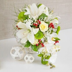 An opulent yet calming floral centerpiece rests on a pristine white tabletop, the kind you might see gracing a wedding reception in a New Barnet restaurant near Victoria Park. The arrangement rises from a low, round white wicker basket, its woven strips criss-crossing to form a subtle pattern that feels both rustic and sophisticated. Dominating the display are several large white lilies, their trumpet-shaped blooms opening with a sense of ceremony, some petals softly recurved to reveal speckles and golden pollen-tipped stamens. At the centre, a single cream-to-pale-yellow rose is nestled securely among the lilies, its layered petals forming a gentle focal point that suggests warmth and affection. Below and to the right, elegant white Phalaenopsis orchids spill gracefully over the edge of the basket, their cascading stems creating a waterfall effect that elongates the composition and adds a contemporary bridal touch. Vibrant greenery fills the spaces between the blooms, from wispy feathery fronds to broader, glossy leaves, giving the design a lush, garden-fresh look, as if freshly gathered from a well-kept New Barnet garden. Bright green pom-pom chrysanthemums introduce a modern, mossy texture, while tiny white sprigs of baby's breath soften the transitions between flowers. Scattered throughout, clusters of deep red hypericum berries provide striking accents, their glossy surfaces catching the light like tiny beads of polished glass. To the left of the basket, two decorative white spheres woven from slim strips of material rest on the tabletop, echoing the basket's craftsmanship and adding sculptural interest. The neutral, textured beige background allows every petal, leaf, and berry to stand out, creating a serene yet celebratory mood that suits weddings, anniversaries, or refined table décor with same-day delivery in and around New Barnet.
