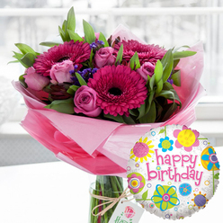A joyful birthday scene unfolds in this image, where a lavish bouquet and playful balloon sit together as if waiting to be carried to a party somewhere in New Barnet. The eye is drawn first to the tall, clear glass vase, filled with fresh water and straight green stems that hint at the bouquet's weight and abundance. Above the vase, the flowers form a full, rounded canopy of colour: bold gerbera daisies in shades of deep fuchsia and bright magenta, their petals radiating like sunbursts around dark, almost black centres, stand proudly at different heights. Between them, layers of blush pink and soft rose-pink roses create a romantic, cloud-like texture, some buds tightly closed, others beginning to spiral open in gentle swirls. Delicate sprigs of deep purple filler flowers peek out here and there, adding depth and a subtle wildflower charm, while glossy green foliage frames the blooms and gives the arrangement a lush, garden-fresh feel, like something gathered near Hadley Green on a sunny morning. The base of the bouquet is wrapped in translucent light pink cellophane, softly crinkled and folded to form a gauzy collar that glows under the natural light. Securely tied around the neck of the vase, a piece of rustic twine holds a small off-white tag printed with a refined rose emblem, signalling a carefully curated gift. In the foreground to the right, a round foil birthday balloon stands out with its white, lightly speckled background, ringed with whimsical, colourful floral and circular motifs in bright yellow, sky blue, lime green, and vivid pink. Across its centre, "happy birthday" is written in lively pink script, echoing the bouquet's palette and capturing the spirit of warm wishes delivered to a doorstep off Wood Street or around East Barnet Road.