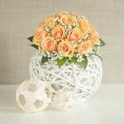 A round, luxurious bouquet of peach roses forms the heart of this image, each rose arranged closely to create a smooth, domed crown of colour that moves from deep apricot at the center of each bloom to softer, creamy peach on the outer petals. Dotted between the roses, sprays of delicate white baby's breath introduce a light, airy texture, like mist settling around the blossoms, while discreet dark green leaves give depth and a touch of garden freshness. The flowers sit inside an eye-catching spherical vase made from fine white wicker strands, carefully woven into an open, geometric lattice design that allows glimpses of stems and shadows within. This sculptural container rests on a spotless white surface, with two smaller woven spheres placed in front: one a pale natural wood tone, the other slightly smaller and lighter, both echoing the same openwork pattern. Together they create a harmonious trio that feels perfectly suited to a contemporary New Barnet living room or a chic celebration near Victoria Park. The background is a soft, neutral beige wall with a faint vertical texture, catching gentle, even light that casts subtle shadows and enhances the intricate lines of the weaving. The overall mood is calm and refined, suggesting a centerpiece created for a wedding, engagement party, or thoughtful gift delivery across New Barnet. The arrangement has the quiet confidence of professional floristry, where every flower head faces outwards and every texture has been chosen to evoke warmth, comfort, and understated elegance.