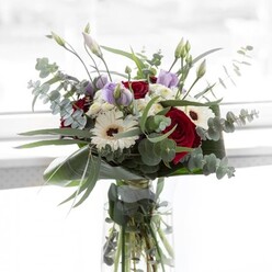 Set against a luminous background reminiscent of a spacious New Barnet home, this meticulously crafted bouquet reflects both contemporary style and classic floral tradition. The arrangement sparkles with velvety red roses at its centre-each petal a testament to enduring affection. Sun-kissed white gerberas, with their striking espresso centers, emerge like radiant stars, imparting an air of celebration. Interspersed among these favourites, soft lilac lisianthus flowers-some still unfolding-whisper promises of new chapters, well-suited for commemorating birthdays or heartfelt milestones in the New Barnet area. Fluffy white filler blooms create a cloud-like softness, cushioning the bolder colours and providing peaceful balance. Eucalyptus, with its cool, silvery-green leaves, arcs gracefully outward, adding both texture and a refreshing woodland aroma reminiscent of nearby Oak Hill Park. Sleek, emerald-hued foliage circles the bouquet's base, grounding the composition with polished sophistication. The glass vase reveals sturdy green stems basking in water, the hallmark of true freshness. Together, these elements echo New Barnet's welcoming spirit-versatile enough to brighten a Barnet High Street shopfront or offer comfort in challenging times. The bouquet's blend of colour, delicate fragrance, and natural textures makes it an expression of gratitude and connection, curated for thoughtful gifting and elegant home décor in this vibrant North London community.