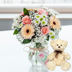 On a bright white tabletop that catches a soft, natural glow, a lovingly styled anniversary gift set takes centre stage, evoking the relaxed charm of a New Barnet living room on a sunny afternoon. A clear glass jar vase holds a compact, rounded bouquet of fresh flowers, their pastel colours blending into a gentle harmony. Pale pink roses unfurl beside peach gerbera daisies with striking dark brown centres, while white chrysanthemums with hints of lime-green in their hearts add purity and light. Airy sprigs of white baby's breath thread between the larger blooms, softening every edge and giving the impression of a light floral mist. The stems show faintly through the glass, a reminder of the flowers' garden beginnings, like something picked from a local garden off Station Road. Around the top of the vase, a length of natural twine is tied in an understated bow, supporting a small rectangular gift tag. The tag is pale and simple, embellished with a pink heart motif and delicate green handwriting that suggests a handcrafted, personal touch. Just to the right, resting on the same gleaming surface, sits a plush cream teddy bear. Its fur appears thick and velvety, inviting a hug; small dark eyes and a chocolate-brown stitched nose give it a sweet, attentive expression. A narrow dark brown satin ribbon is neatly knotted at its neck, adding a classic finish. The softly blurred, light-filled background ensures all focus remains on this intimate pairing-a bouquet and teddy bear that together whisper affection, reassurance, and celebration, ideal for anniversaries, new arrivals, or heartfelt get-well wishes in and around New Barnet.