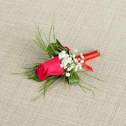 A delicately styled boutonniere rests at a gentle angle on a pale beige, linen-like fabric, its textured weave recalling the relaxed, homely feel of New Barnet houses near Victoria Park. At the centre of the design, a single rosebud in vivid fuchsia, almost leaning towards deep magenta, remains tightly furled, suggesting anticipation and new beginnings. The petals are velvety and richly coloured, with smooth edges that catch the bright overhead light, giving the bloom a subtle satin sheen. Encircling this focal rose, small tufts of white baby's breath cluster like tiny stars, their miniature blossoms adding lightness and softness to the composition. Slender, bright green, feathery foliage radiates outward behind the flowers, the fine fronds echoing the look of asparagus fern and lending the boutonniere a natural, slightly whimsical silhouette, as if freshly gathered from a garden just off East Barnet Road. Nearer the base, a few broader, darker green leaves provide balance and depth, anchoring the airy textures above. The stems of the rose, baby's breath, and greenery are carefully aligned and bound together with a narrow red satin ribbon, wound snugly around the base and finished with a small, discreet detail that hints at a florist's skilled, practised hands. The neutral, softly lit background allows the bold pink, crisp white, and layered greens to stand out clearly, creating an image that feels both romantic and composed. It evokes the quiet excitement of fastening a buttonhole to a suit jacket before a wedding ceremony or prom night in New Barnet, a detail that brings colour, fragrance, and a touch of tradition to a special occasion.