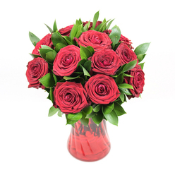 In this image, a classic, almost theatrical bouquet of approximately twelve to fifteen deep red roses stands proudly in a translucent red glass vase, lit evenly against a spotless white backdrop. Each rose is at its peak, with layers of velvety petals unfurling in perfect spirals, creating a sense of depth you could almost brush with your fingertips. The red tones are intense and saturated, somewhere between ruby and wine, and they dominate the scene, echoing the kind of passionate gesture you might send across New Barnet for an anniversary or a private celebration at home near High Barnet station. Generous clusters of dark, glossy green leaves, likely ruscus or similar foliage, weave between the blooms, giving structure and contrast. The leaves appear fresh and supple, catching small highlights along their edges where the studio light touches them, which helps to separate each rose head and prevent the bouquet from feeling flat. The vase has a gently curving silhouette that widens at the top, its subtly ribbed or wave-like texture bending the light into ripples of red, almost like reflections on the surface of Pymmes Brook after rain. Because the glass is translucent rather than fully opaque, you sense the stems descending into the water, anchoring the arrangement with a quiet naturalism even though they are not the focus. The stark white background strips away any distractions, so all attention goes to the harmonious interplay of rich red petals, cool glass, and vibrant green foliage. This is the kind of rose bouquet a skilled local florist would create when someone wants to express undiluted emotion-romantic, confident, and timeless.