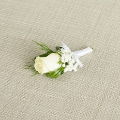 A classic wedding boutonniere rests at a gentle diagonal on a pale beige, linen-like fabric, the weave of the material forming a subtle crosshatch that frames the flowers without drawing attention away from them. At the heart of this refined piece is a single white rosebud, creamy and velvety, its petals wrapped closely together with just the slightest curl at the edges, as if moments away from unfurling fully. The bloom has a soft, luminous quality, as though caught in calm daylight streaming through a New Barnet townhouse window before the bridal party sets off to the ceremony. Tucked around and just behind the rose are tiny sprays of white baby's breath, each delicate floret creating a misty, cloud-like ring that makes the rose appear even more pristine. The filler flowers look airy and weightless, yet they provide a gentle fullness that balances the simple elegance of the single stem. Slender, dark green leaves and a couple of fine, ferny fronds provide contrast, their smooth and feathery textures playing against the satin finish of the rose petals. The foliage is arranged so the greens echo the natural curve of a lapel, suggesting how it will sit against a tailored suit at a New Barnet church or registry office near the High Road. The stems are tightly gathered and expertly wrapped in glossy white satin ribbon, the wrap clean and even, ending in a small, neatly tied bow that adds a touch of handcrafted charm. The bow's loops sit just below the flower heads, giving a clear visual cue where a pin would secure it in place. Soft, diffused lighting gently brings out every contour of the rose and the tiny shapes of the baby's breath, casting only the faintest shadows and preserving a calm, romantic mood. This boutonniere feels perfectly suited to a groom or groomsman stepping out onto the leafy streets around New Barnet for a day of quiet celebration and heartfelt promises.