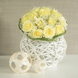 On a pristine white tabletop, a lush, dome-shaped bouquet of soft yellow roses takes center stage, each rose carefully placed so that the petals touch and overlap, forming a seamless, buttery carpet of bloom. Between these roses, fine sprays of white baby's breath weave gently in and out, their tiny blossoms catching the light and giving the arrangement a cloud-like softness that feels perfectly suited to a wedding breakfast near New Barnet Station or an intimate celebration at home. A slim ring of rich green foliage is wrapped snugly around the base of the flowers, like a natural ribbon, tidily finishing the shape and highlighting the florist's attention to structure and proportion. The bouquet rests in a distinctive white woven vase, its body formed from crisscrossing strands that mimic modern wicker, leaving small openings that let the light dance through. This airy design, reminiscent of a contemporary basket you might find in a stylish boutique off Station Road, lends the centerpiece an elegant, modern character while keeping the mood warm and welcoming. Beside the vase sit two decorative balls made from pale, natural-looking rattan or wood strips, their open, spherical frames echoing the same geometric weave and creating a gentle visual dialogue between all the elements. The background is a soft, neutral beige, blurred just enough to suggest depth without distraction, much like looking past a beautifully dressed wedding table towards the calm walls of a New Barnet venue. Together, the creamy roses, feathery gypsophila, and textured white decor create a scene that feels serene, romantic, and thoughtfully crafted for a special table arrangement.
