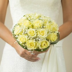 Bathed in the gentle glow of late morning, this striking bridal bouquet is showcased in the hands of a bride poised against the intricate white fabric of her wedding gown, somewhere along the quiet streets of New Barnet. The floral focal point is a symphony of closely nestled pale yellow roses, their velvety petals spiraling outward in soft shades of butter and lemon, each blossom opening just so to evoke a sense of new beginnings near East Barnet Village. Threaded throughout the roses are subtle tufts of airy white gypsophila, or baby's breath, their tiny blossoms suspended like gentle morning mist-injecting an ethereal charm into the lush ensemble. The bouquet is further defined by the careful addition of thin spiral lines of glossy dark green foliage, which curves beneath the blooms in a graceful sweep, offering a contemporary twist rooted in skilled local floristry. The bride's hands are perfectly manicured, her nails in a soft blush hue, and a simple gold band glints on her ring finger, underscoring the intimacy and meaning of the moment. The softly blurred background ensures focus rests on the bouquet's radiant colors and rich textures. Ideal for wedding dreamers across New Barnet, this artful composition captures the essence of local floral tradition, romance, and celebration.