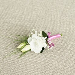 An elegant white lisianthus boutonniere rests diagonally on a softly textured beige fabric, the surface reminiscent of finely woven linen you might see dressing tables in New Barnet's period homes. The central flower is fully open, a pristine white bloom with gently ruffled petals that curl and layer into a rounded, romantic form, inviting the eye to trace each soft fold. Nestled close beside it are two slender lisianthus buds in a creamy, pale green shade, their tips still closed, capturing that tender moment of anticipation so fitting for a wedding morning or formal celebration. Around these focal blooms, tiny stars of white gypsophila drift like a delicate mist, filling the gaps with an airy, almost ethereal lightness, just as a skilled florist would arrange them for a groom preparing near New Barnet station or along the High Road. Slim, grass-like strands of greenery arc out toward the lower left, adding graceful lines and a sense of movement, while a single darker green leaf supports the main bloom from behind, grounding the composition with depth and contrast. At the base, the stems are neatly bound in a bold band of fuchsia pink satin ribbon, tied into a tidy, cheerful bow that injects a flash of modern colour into the classic ivory-and-green scheme. The gentle, even lighting showcases each texture-from the silky petals to the satin sheen of the ribbon-creating a refined, handcrafted accessory perfect for lapels at local New Barnet weddings, proms, or black-tie gatherings, and reflecting the care and artistry of experienced neighbourhood florists.