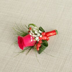 A handcrafted boutonniere lies diagonally across a softly woven light beige fabric, its subtle texture evoking the calm, neutral tones of a well-dressed suit or wedding table linen. The focal flower is a single fuchsia-pink rose, its centre tightly coiled and its outer petals gently unfurling, with a plush, velvety quality that suggests it has just been conditioned by an expert florist. Tiny white baby's breath flowers cluster around the rose like a cloud, their numerous little blooms adding a light, airy counterpoint to the bold pink, much like the soft mist that sometimes lingers over the green spaces of New Barnet in early morning. Tucked among these white sprigs, glossy red hypericum berries gleam, their rounded shapes and vivid colour introducing a festive spark that feels perfect for a winter wedding or Christmas party near Barnet High Street. Threading through the composition are fine, feathery stems of fern-like greenery that spill outward with gentle movement, while broader dark green leaves have been carefully curled and shaped to frame the rose, creating a balanced, architectural look. The stems are tightly wrapped in a smooth red satin ribbon, finished with a small, tidy bow that sits snugly against the fabric and gives a sense of tailored polish. Viewed from slightly above, the image reveals every layer-the interplay of greens, the crisp whites, the shining berries, and the intense rose at the centre-forming a small yet expressive piece that would sit proudly on the lapel of a groom, groomsman, or prom guest heading out from New Barnet to celebrate a cherished occasion.