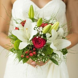 The beauty and emotion of a wedding morning in New Barnet come alive in this detailed close-up: a bride in a strapless white gown, softly textured, holding a bouquet that feels like a joyous secret whispered along local high streets and the gentle pathways of Oakhill Park. The bouquet's heart is sculpted from pure white oriental lilies, their petals wide and gleaming as they open toward the future, each golden stamen catching the soft light. Nestled with care are passionate red roses, their tightly furled petals embodying both intensity and quiet confidence-symbols of romance cherished in every New Barnet love story. Sprigs of baby's breath drift among the flowers like fine lace, airy and light-while the inclusion of unopened green lily buds hints at beginnings yet to blossom. Scarlet hypericum berries and quirky green orbs add playful notes of colour and texture, reminiscent of fresh finds from the neighbourhood flower markets. Foliage spills from the base-some fern-like, some broad and verdant-granting fullness and natural depth to the composition. The bride's hands, gently enclosing the stems, express the moment's significance: a bouquet that's not just decoration, but a talisman of hope, commitment, and local artistry. Such an arrangement, meticulously crafted for a New Barnet celebration, is destined to become a treasured memory woven into the fabric of a couple's journey together.