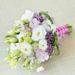 A radiant, hand-tied bouquet lies gently on textured neutral linen, capturing the spirit of a New Barnet bridal morning near the High Road. Creamy white lisianthus, velvety and open, share quiet space with tightly furled lime-green buds, their juxtaposition a subtle nod to both promise and bloom. Soft lavender-pink stock provides a cloud of fragrance and colour, bringing to mind the gentle transition from spring to summer across North London gardens. Around the main blooms, deep purple allium stars project outward, their geometric precision paired with a wild, celebratory energy. Slender green bear grass loops add a sense of movement, like the gentle sway of grass in Victoria Recreation Ground. The bouquet's stems are expertly bound with vivid fuchsia satin ribbon, accented by a string of white pearls-a sophisticated finish that sparkles in soft daylight. Intermingled greens and textured filler frame the arrangement, each element chosen to add depth and variety while maintaining harmony. The result is a bouquet that feels alive and cherished, crafted for those moments of connection and joy celebrated across New Barnet. Whether for weddings, showers, or meaningful gatherings, this arrangement embodies optimism and artistry, reflecting the care and creativity of New Barnet's dedicated florists.