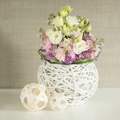Nestled on a pristine white surface beside the inviting streets of New Barnet, this floral arrangement is a subtle masterpiece, expertly composed by a local florist. The bouquet overflows with creamy lisianthus and pale yellow blooms, whose soft, ruffled petals dance lightly around upright pink matthiola, infusing the display with both volume and grace. Deep purple aster-like blossoms create a vivid bed at the base, their tiny star shapes adding delightful contrast beneath the pastel canopy. Fresh green foliage forms a natural border, wrapping the flowers in a vibrant embrace. The bouquet is presented in a distinctive, round white vase, its openwork of entwined painted rattan or branches creating an architectural yet delicate finish-reminiscent of thoughtful design inspired by the unique character of New Barnet. Flanking the vase are two spherical accents woven from pale wood, their intricate latticework echoing the vase's textural charm. Soft, filtered light glides across the arrangement, casting gentle shadows and illuminating every nuanced fold and hue. Against a backdrop of gently textured beige reminiscent of local café interiors, the entire scene radiates tranquility and warmth-a visual serenade to special celebrations, whether marking a wedding at nearby Monken Hadley or brightening a family gathering in northern Barnet. Every detail speaks to seasoned expertise and heartfelt care, capturing the spirit of community and creativity that defines the area.