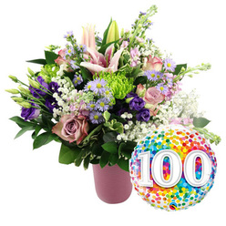 Set against a clean, uncluttered white background, this image captures a joyful 100th celebration bouquet that feels ready to be delivered to a New Barnet home for a once-in-a-lifetime milestone. The arrangement is gathered in a contemporary ribbed vase in soft matte pink, its cylindrical form gently anchoring a lush, almost cloud-like mass of flowers that billow above. At the crown of the bouquet, light pink Oriental lilies stretch their elegant petals outward, some fully open to reveal long, graceful stamens dusted with pollen, others still in poised buds that promise future blooms. Around them, layers of pale blush roses and deeper dusty rose-toned roses nestle closely together, their overlapping petals creating a sense of depth and quiet luxury. Threaded through the design are rich purple lisianthus flowers with satiny petals, interspersed with airy clusters of tiny lavender asters, each one finished with a bright yellow centre that glows softly against the cooler tones. Pops of lime green button chrysanthemums add an energetic twist, their tight, domed heads echoing the playful feel of a party. Wisps of white baby's breath drift throughout the bouquet, filling small gaps and giving the entire arrangement a floating, light-as-air effect, while a mix of dark and mid-green foliage frames the flowers and creates a fresh, garden-picked impression. To the lower right of the vase, slightly overlapping the bouquet, a glossy round foil balloon shimmers with multi-coloured confetti dots in vivid shades of red, orange, yellow, green, and blue. At its centre, large white numerals read 100 with a subtle three-dimensional shading, instantly signalling a centenary birthday or anniversary celebration somewhere in the heart of New Barnet.
