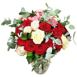 A lavish rose arrangement, captured against a bright white background, shows off a carefully composed blend of colour and texture that feels both classic and fresh, like a favourite walk through New Barnet's quiet residential streets after rain. The bouquet is dominated by deep red and scarlet roses, their velvety petals opening layer by layer, some standing tall in bud, others fully unfurled, creating a rich sense of movement and romance. These passionate tones are softened by clusters of creamy ivory and pure white roses, with the occasional bloom lightly brushed at the tips with the palest pink, echoing the gentle warmth of a late afternoon sky. Nestled throughout, tiny bright pink filler flowers, similar to small carnations or berry-like sprays, punctuate the design, giving a playful contrast and a touch of brightness around the more formal roses. Flowing through and around the flowers are plenty of rounded eucalyptus leaves in muted blue-green, their slightly matte surfaces and relaxed curves adding a modern, botanical character, along with the suggestion of a cool, calming aroma. Additional dark green foliage and a hint of silvery-green leaves provide structure, so the bouquet forms a generous, rounded dome that looks lush from every angle. The clear glass vase, with its slightly bulbous body and narrower neck, cradles the stems in visible, clean water, giving the composition stability and a sense of transparency and honesty. Styled like this, the bouquet could sit proudly in a New Barnet living room or restaurant, perfect for romantic celebrations, proposals, or simply bringing a touch of refined beauty into an everyday local home.