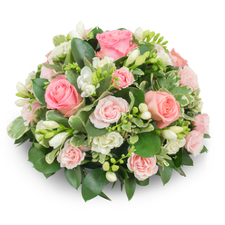 Viewed head-on, this compact, rounded floral cushion conveys a composed and comforting mood: dominant, fully opened pink roses-ranging from creamy blush to a slightly deeper coral-pink-are positioned to create a rhythmic, domed surface that holds the eye. Between these focal roses, clusters of pristine white freesia offer vertical punctuation; some blooms are open and delicate, showing pale yellow centers, while others remain in snug bud form, suggesting the arrangement will breathe and evolve over time. Interspersed spray roses and small white filler blooms provide soft layering, their finer petals adding a delicate counterpoint to the larger rose faces. The greenery is deliberately lush-dark glossy leaves create a dense, supportive matrix while variegated, cream-edged foliage trims the silhouette and adds a whisper of light. No stems are visible, reinforcing the formal, finished effect of a florist's cushion bouquet. Natural daylight brings out the satin-like texture of the rose petals and the subtle translucence of freesia bells, bathing the composition in a soothing, neutral glow. As a sympathetic tribute or refined gift, this piece is crafted to comfort: its palette and proportions foster reflection and tenderness. Locally minded and hand-finished, this style is often chosen for memorial services in and around New Barnet, where neighbours appreciate understated elegance and floral messages that convey condolence with grace.