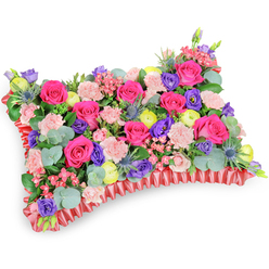 From a slightly raised, angled viewpoint the image presents a meticulously crafted floral cushion, a rectangular pillow of blooms that reads as both art and tribute. Hot pink roses dominate the surface - their petals velvety and layered, some tight as buds, others opening to reveal warm depths - while pale pink carnations knit between them, lending a delicate, frilly texture across the top. Scattered throughout are rich purple lisianthus (eustoma) whose deeper tones add contrast and gentle shadow, and bright lemon-yellow ranunculus that inject sunnier highlights reminiscent of late spring sunshine. Eryngium thistles, with their steely blue spikes, provide an unexpected architectural edge against the softer florals, and generous sprigs of rounded eucalyptus leaves, silvery-green and aromatic, trail through the design to give cooling contrast and graceful movement. The cushion rests on a decorative base finished with a pleated, deep-red satin ribbon that ruffles around the perimeter, framing the arrangement like a soft, elegant trim and reinforcing the pillow silhouette traditionally used as a sympathy or memorial tribute. The careful placement of textures - smooth rose petals, frilled carnation edges, spiky thistle points and rounded foliage - shows experienced craftsmanship and an eye for balance. The overall mood is one of tender remembrance rather than mourning; it celebrates life with vibrant fuchsia, violet, lemon and blush tones that comfort more than they grieve. As local florists serving New Barnet and neighbouring streets around Station Road, Flowers New Barnet crafts pieces like this for funerals, memorial services or quiet family tributes in nearby Oak Hill Park or at local chapels.