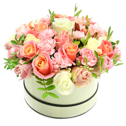 Presented in a round, cream hatbox trimmed with two slim dark bands that frame its clean silhouette, this lush floral design feels both luxurious and intimately familiar. The arrangement forms a dense, domed cluster of fresh blooms, dominated by generously sized roses that graduate in colour from warm coral-peach and apricot at their centers to softer blush pinks and pristine ivory at their edges, capturing a soft ombre that reads as romantic and optimistic. Interspersed among the larger roses are flashes of light-pink, ruffled carnations whose frilly texture creates gentle contrast and fills the bouquet with tender detail. Scattered sprigs of vivid green foliage, with small, elongated leaves, breathe life into the composition and add a crisp, natural counterpoint to the velvety petals. Against a stark white background and bathed in soft, natural light, the petals' subtle sheen and layered tones become especially visible, inviting the eye to travel across each bloom. The overall sensory impression is of quiet elegance - a soft, powdery fragrance, plush textures, and the reassuring weight of a thoughtfully arranged gift. As a local florist serving the New Barnet community, Flowers New Barnet arranges these hatbox bouquets to mark birthdays, anniversaries, Valentine's gestures, or simply to say thank you; many customers choose this style for deliveries around New Barnet Station, Monken Hadley Common, and nearby Oakleigh Park because it reads as both celebratory and comfortingly refined.