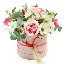 In this photograph, a chic, pastel pink hatbox brimming with flowers is set against a pristine white backdrop, allowing every petal and leaf to stand out in gentle detail. The box itself is cylindrical and softly matte, tied with a slender coral-red satin ribbon that wraps around the middle and gathers into a precise bow at the front. From the bow hangs a cream gift tag printed with green script, its neutral tones echoing the natural palette of the bouquet and inviting a handwritten note before delivery around New Barnet. Above the rim of the box rises a lush, rounded dome of blooms, full and carefully structured yet relaxed enough to feel natural. A striking bi-colour rose in pale pink with a richer pink swirl at its centre anchors the design, its velvety petals opening generously like a focal point one might pause to admire on a stroll past local shopfronts. Surrounding this rose, white daisy-style chrysanthemums with bright green centres add brightness and freshness, their tidy petals radiating outward like tiny suns. Slender white alstroemeria, dusted inside with fine speckles, weave between them, and deeper pink flowers-perhaps spray roses or lisianthus-cluster in pockets of intense colour, giving depth and rhythm. Silvery eucalyptus leaves arc delicately from the mass of blooms, their rounded shapes softening the edges, while darker greenery and small budding stems provide texture and volume. The arrangement feels romantic, polished, and quietly joyful, perfectly suited to occasions such as Mother's Day, anniversaries, or a thoughtful surprise to a friend near East Barnet or Monken Hadley. The overall impression is of a refined, handcrafted floral gift that captures a moment of celebration in a calm, contemporary style.