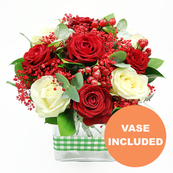 Against a bright, uncluttered white backdrop, a festive rose arrangement is showcased as if on a spotless display stand in a New Barnet studio. The composition centres on generous red roses and creamy white roses, each blossom fully open, revealing intricate whorls of petals that create a luxurious, almost velvety surface. These main flowers are threaded through with sprays of tiny red filler blooms that resemble coloured gypsophila, giving the bouquet a fine, cloudlike texture and a joyful, confetti-style effect. Nestled throughout, glossy red hypericum berries catch the light with a soft sheen, their rounded forms adding a sculptural contrast to the softness of the petals. Surrounding the roses, a fresh mix of foliage weaves in and out: silvery-green eucalyptus with its slightly rounded leaves, and deeper green foliage that fans outward, giving the arrangement both structure and movement, like a gentle breeze passing through the trees by Tudor Park. Everything is gathered into a modern, rectangular clear glass vase, allowing you to see the cleanly cut stems aligned inside, a detail that speaks to the care and precision of a professional florist. A green and white gingham ribbon wraps horizontally around the centre of the vase, its checked pattern bringing a homely, country-inspired character that would suit a New Barnet kitchen table or living room windowsill. In the bottom right corner, an orange circular graphic clearly declares "VASE INCLUDED" in bold white letters, emphasising that the piece is ready to place the moment it arrives, perfect for same-day delivery surprises for birthdays, anniversaries, or cosy seasonal celebrations in and around New Barnet.