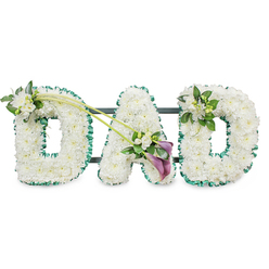 Elegant funeral flower tribute spelling out "DAD" in large upright letters, designed in a timeless style that would feel at home in any New Barnet church or local crematorium chapel. Each letter is carefully built on a sturdy, dark-backed frame, then completely carpeted with tightly clustered white chrysanthemums or similar small white flowers, creating a dense, velvety texture with no gaps between the blooms. Around the perimeter of the letters runs a vivid emerald-green satin ribbon, softly ruffled and pleated, which gives a crisp outline and hints at the fresh greenery found in nearby New Barnet parks and gardens. Subtle floral sprays soften the form: on the first "D", a small cluster of white freesia and glossy green leaves sits gracefully on the upper left curve, while a similar, slightly smaller accent decorates the upper right of the final "D". From the top of the first "D", a pair of slender green stems arcs gently toward the central "A", guiding the gaze to a more dramatic focal arrangement. Here, two deep purple calla lilies nestle among white freesia and foliage, their smooth, sculptural trumpets introducing a note of solemn beauty and deeper colour. The tribute is set against a bright white background, highlighting every petal, ribbon fold, and curve of the lettering. The composition conveys quiet strength, respect, and heartfelt loss, an intimate memorial piece that allows family members in New Barnet to honour their father with a personal word formed entirely from fresh flowers.