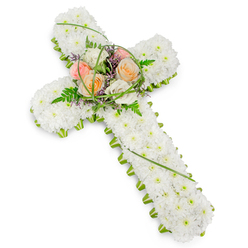 A traditional Christian cross made entirely of flowers appears here against a bright white background, its form clear and unwavering, as though prepared for a thoughtful farewell in New Barnet. The body of the cross is fully covered in closely packed white chrysanthemums, or similar small white blooms, so that no gaps remain, just a seamless, cloud-like surface of petals. This dense covering creates a sense of softness and rest, inviting the eye to trace the contours of the design. Around the perimeter, a neat trim of vivid green foliage outlines each edge, from the top arm to the base, subtly framing the white and emphasising the cross shape with crisp definition. At the very centre, where the vertical and horizontal stems meet, a delicate focal spray brings warmth and quiet colour. Several roses, fully open and velvety, appear in a gentle spectrum of peach, pale pink, and creamy ivory, their tones blending harmoniously rather than competing. Fern fronds curl gently around them, while fine strands of bright green bear grass loop and arch in soft lines. Tiny lavender-purple filler blossoms peek through the greenery, giving a hint of depth and shadow. The lighting feels natural, as if this tribute were resting in a peaceful chapel near New Barnet High Street, ready for a funeral, memorial service, or final farewell. The overall impression is one of composed elegance and heartfelt respect, a floral cross that conveys love, solace, and faith for families seeking a meaningful sympathy arrangement in the New Barnet area.