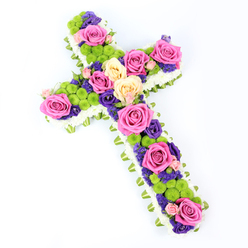 Set against a spotless white background, this cross-shaped funeral tribute appears almost to float, its colours glowing softly as though lit by gentle chapel light. The structure of the cross is clearly defined, with straight, even arms and a balanced proportion that feels both traditional and reassuring, the kind of design often chosen by families in New Barnet when they want flowers to speak quietly but clearly of faith and love. The base of the cross is edged in crisp white spray chrysanthemums, their tiny petals forming a dense, cloud-like cushion that outlines every curve and corner. Just beneath this white halo, a discreet band of folded light green ribbon or foliage offers a fresh, verdant edge, as if a whisper of Oak Hill Park's greenery had been woven in for comfort. Within this frame, the interior bursts into a tapestry of colour: large roses in hot pink and creamy pale yellow unfurl their layered petals, each bloom carefully spaced to guide the eye along the length of the cross. Nestled between them are rounded lime-green button chrysanthemums, their playful pom-pom shapes bringing a modern touch, while clusters of deep purple blooms-likely lisianthus or statice-thread through the design, adding shadows of rich colour and a contemplative tone. The flowers look freshly arranged, petals unblemished and faces wide open, as though prepared that morning for a funeral or memorial service in New Barnet. The overall effect is one of gentle hope and dignified remembrance, a floral cross that offers solace while quietly celebrating a life well lived.