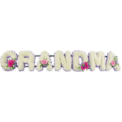 This image shows a beautifully composed funeral flower tribute spelling the word "GRANDMA" in large, upright letters, the sort you might see resting beside the chapel at New Southgate Cemetery or a small service in New Barnet. Each letter is completely covered in soft, cloud-like white chrysanthemums, closely placed so that no base material is visible, giving the impression of a continuous, cushioned surface. The petals are full and slightly ruffled, catching the light with a gentle sheen that adds depth and movement to the design. Running around the edge of every letter is a slim band of lilac ribbon, neatly folded and pinned, its pastel tone lending an air of serenity and quiet grace. Three focal posies break up the white and lilac: one at the lower curve of the "G", another embracing the middle "A", and a final cluster resting on the last "A". These mini bouquets feature cheerful pink gerbera daisies and soft pink roses, interwoven with fresh greenery that curls and fans out, suggesting growth, continuity, and renewal. The greens are a natural, garden-fresh shade, like foliage you might see in a well-kept front garden off Lyonsdown Road. The lighting is soft and even, without harsh shadows, allowing every flower head and ribbon edge to be clearly defined. Together, these elements create a tribute that feels both tender and composed, a floral message of love and remembrance for a much-loved grandmother, thoughtfully arranged by skilled hands familiar with the needs of local New Barnet families during times of farewell.