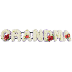 A tender funeral flower arrangement spells out the word "GRANDMA" in large capital letters, stretching horizontally across a pure white backdrop that keeps the focus entirely on the flowers. Each letter is formed from closely set white cushion chrysanthemums, their compact, starry blooms creating a rich, cushion-like surface that suggests softness and comfort. Around the outer line of every letter runs a subtle trace of pale purple ribbon or backing, just visible between the petals, giving the design a gentle, lavender haze and a touch of refined elegance. Bold red accents break through the white, placed thoughtfully in only a few spots so they feel meaningful rather than overwhelming. On the "G", a cluster of deep red roses is gathered with a distinctive variegated red-and-cream calla-style flower, tucked neatly into the curve of the letter. The first "A" is finished with a single heart-shaped red anthurium, its waxy, polished texture catching the light. In the "M", more velvety red roses and another anthurium nestle into the blooms, while the final "A" carries one last striking anthurium, completing a subtle rhythm from start to end. A dark green metal frame or stand supports the letters, tilting them slightly forward as if to present the tribute to those gathered. This carefully balanced sympathy design would sit beautifully beside a coffin in a New Barnet chapel, or at a crematorium service near Barnet High Street, offering a personal, loving farewell from grandchildren and family members who want their message of "GRANDMA" to be seen and felt at a glance.