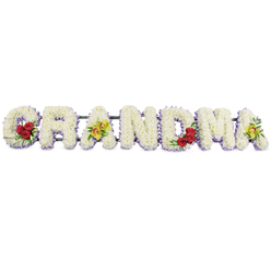 The image shows a lovingly crafted floral tribute spelling out GRANDMA in striking block capitals, presented in a straight line against a bright, uncluttered white background. Each letter is formed from a dense bed of small white chrysanthemums or similar button blooms, tightly packed so that no gaps in the petals are visible, giving the whole word a smooth, cushioned appearance. Along the lower edges of the letters runs a delicate border of soft lavender flowers, providing a gentle contrast and helping each shape stand out with quiet clarity. At the far left, the G is embellished on its lower curve with a cluster of three deep red roses, their rich colour and glossy greenery adding depth and emotion. The first A carries two elegant yellow-green orchids on its central bar, their pale petals and speckled centres introducing a more unusual, graceful note. In the middle of the tribute, the R, N and D are kept purely white with the lavender edging, allowing the eye to rest on the calm simplicity of the design. Moving right, the M echoes the opening letter with another trio of velvety red roses nestled into its lower right section, while the last A once again features a pair of green orchids, visually balancing the tribute from end to end. A dark green or black metal frame supports the letters from behind, almost invisible against the white setting but essential for stability during transport and display. The overall effect is gentle, dignified and full of love, a fitting floral farewell from a New Barnet family saying goodbye to a cherished grandmother at a local church or graveside service.