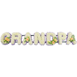 A serene floral name tribute spells out "GRANDPA" in large block letters, all arranged in a straight horizontal line as if resting on an invisible shelf. Each letter is generously filled with small, pristine white chrysanthemums or similar dainty blooms, packed so closely that they create a plush, cushion-like surface, soft and rounded at the edges. Tracing the outline of every character is a slim border of pale purple or lilac flowers, introducing a gentle wash of colour that clearly defines the contours of the G, R, A, N, D, P, and final A. These lavender tones bring a sense of calm and quiet reflection, ideal for a funeral or remembrance gathering in and around New Barnet. Within the structure of the arrangement, select letters carry delicate focal sprays: the 'G' and 'D' each hold a cluster of peach roses, some just opening, surrounded by polished green leaves and slim white lily buds, while the first and last 'A' are each adorned with a single, perfectly placed peach rose and foliage. The interplay of white, peach, and lilac evokes feelings of peace, gratitude, and enduring affection for a much-loved grandfather. Supported by a dark, low-profile frame, the piece appears sturdy yet unobtrusive, designed to stand gracefully beside a coffin or near the front of a chapel. The crisp white background in the image enhances every petal and subtle colour variation, much like the way careful lighting in a local New Barnet funeral home might highlight the tribute during a quiet moment of farewell.