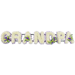 The image shows a carefully crafted funeral flower tribute that spells out GRANDPA in bold, upright block letters, set out in a clean horizontal row against a bright white background, as though placed on trestles at the front of a service. Every letter is formed from a dense carpet of small white flowers, most likely fresh chrysanthemums or carnations, packed tightly together so no gaps or stems interrupt the smooth, velvety surface. The white blooms create an impression of serenity and purity, their rounded petals giving each letter a pillowed softness that speaks of comfort and quiet remembrance. Threaded through this sea of white are small yet meaningful clusters of rich purple and lilac blossoms, accompanied by fresh green foliage. These accents are positioned with intention: tucked into the curl of the 'G', in the central triangle of the first 'A', along the upright of the 'D', within the loop of the 'P', and resting delicately near the final 'A'. This careful placement draws the eye gently along the name, much like pausing at shared memories of a grandfather's stories, kindness, and guidance. The letters rest on a sturdy, dark green frame that runs invisibly beneath them, ensuring everything stays aligned and secure for transport to churches, homes, or chapels of rest throughout New Barnet and the surrounding residential streets. The overall feeling is one of deep affection and respect, a personalised floral message that would sit gracefully beside a coffin or memorial photograph, offering a comforting focal point for grieving family and friends saying goodbye to Grandpa in the New Barnet community.