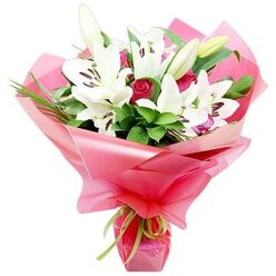 An artfully composed bouquet of white Oriental lilies and deep red roses stands out crisply against a clean white background, showcasing the skill of a local New Barnet florist. The lilies are the stars of the design: large, trumpet-shaped blooms with pristine white petals that flare outwards, each bloom revealing a cluster of dark reddish-brown anthers poised on slender filaments, dusted with fine pollen. Around these open flowers, several plump lily buds, shaded in soft green and cream, reach upwards, suggesting freshness and the promise of new blossoms over the coming days. Intertwined among the lilies are rich red roses in various stages of opening, from tightly furled buds with a velvety sheen to more relaxed flowers beginning to unfurl their layered petals, creating a romantic counterpoint in both colour and texture. The composition is framed by a generous amount of greenery: deep green, slightly waxy leaves and slimmer, grass-like blades that arc gently, giving the bouquet height and movement. All the stems are gathered into a tight hand-tied cluster and wrapped in layers of glossy, bright pink cellophane or floral paper, which forms soft, billowing folds around the flowers and reflects light like a festive gift bag carried down East Barnet Road. At the base, a slender lime green ribbon is secured in a neat bow, the cool green hue playing off the foliage and adding a modern twist. The overall mood is joyful and affectionate, ideal for New Barnet flower delivery when marking birthdays, anniversaries, graduations, or heartfelt thank-you moments.