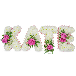 A lovingly crafted sympathy tribute spells the name "KATE" in large, three-dimensional letters, each one composed of a dense blanket of small, pristine white chrysanthemums that read as a soft, cloud-like canvas. A delicate pale pink satin ribbon outlines the inner edges of every letter, creating a gentle contrast and a quietly feminine silhouette. Hot pink roses provide vivid focal points - two opened roses adorn the K, a single striking rose nestles in the lower curve of the A, one intense bloom sits at the base of the T, and a pair of roses accent the right side of the E - their silken petals catching the light and releasing a faint, comforting rose scent. Interwoven green elements - ferny fronds, ruscus sprigs and variegated leaves - lend structure and a fresh, garden-like breath between the floral clusters. Presented as a freestanding display against a clean white backdrop, subtle shadows beneath the letters give the tribute dimension and dignity, ideal for memorials or celebrations of life at local venues, churches, or funeral homes in New Barnet and nearby High Barnet. Prepared by Flowers New Barnet with the same care we bring to local deliveries, this piece balances tenderness and craftsmanship, offering a personal, heartfelt expression of remembrance that resonates with families in the neighbourhood and beyond.