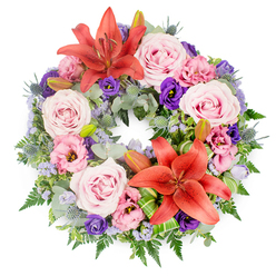 A richly detailed sympathy wreath lies against a pure white background, its circular form completely filled with flowers and foliage so that no bare frame shows through. At first glance the eye is drawn to two bold Asiatic lilies in a warm coral-orange, their petals wide open and gently curled at the tips, one anchoring the upper section of the ring and the other adding balance near the lower right. Around them, numerous roses in soft blush and lavender-pink tones nestle closely together, their layered, velvety petals suggesting tenderness and affection. Between the roses, ruffled lisianthus blooms appear in delicate pinks and deeper purples, their cup-shaped flowers adding movement and texture as they tilt in different directions. Small, spiky blue eryngium thistles punctuate the softer blossoms, their cool steel-blue heads and jagged collars providing a subtle, modern edge that contrasts beautifully with the pastels. Fine sprays of tiny lavender-blue filler flowers, likely statice or limonium, weave through the composition like a light haze, softening edges and creating a sense of depth. The base of the wreath is a lush mix of greenery: feathery ferns, smooth, rounded leaves in muted blue-green, and brighter green foliage that adds freshness. Artfully folded accent leaves cradle some of the blooms, particularly near the lower lily, showing the hand of an experienced florist. A few closed lily buds peek from the foliage, hinting at blossoms that would open in the days following the service. Created in the calm, residential surroundings of New Barnet and suitable for same-day delivery to local churches, crematoriums, or homes, this wreath expresses comfort and remembrance with quiet elegance.