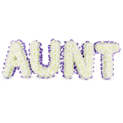 As a local New Barnet florist I describe this tribute as a crafted message of love shaped entirely from pure white spray chrysanthemums that spell "AUNT" in bold uppercase letters. Each character is densely filled with hundreds of small, ruffled chrysanthemum florets whose delicate petals form a cushiony surface; here and there pale yellow eye-centres warm the whiteness like tiny sunlit details. A deep purple satin ribbon, pleated and gently ruffled, outlines each letter, hugging the frames with a soft, tactile edge that throws a modest highlight against the white flowers. Light from a nearby window at times catches the ribbon's sheen and sets a quiet contrast, while faint grey shadows beneath each letter clarify the three-dimensional build and steady structure. You can almost sense the clean, green-tinged fragrance of freshly cut chrysanthemums rising from the arrangement-subtle, comforting, and natural. Designed with the calm restraint appropriate for a funeral chapel, family service, or a remembered corner at home, the combination of white blooms and lavender-edge ribbon conveys dignity and loving remembrance rather than sombreness. At Flowers New Barnet we arrange these personalised letter tributes for services near New Barnet Station, around Station Road and the Victoria Recreation Ground, and they are available for local delivery to churches and private addresses across the area. The piece reads as both a naming of loss and an offering of quiet gratitude, made with steady hands and neighbourhood care.
