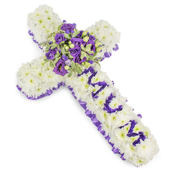 A dignified Christian cross crafted from an abundance of pristine white chrysanthemum blooms, each petal layered to create a cushioned, continuous surface that reads as serenity and reverence. The cross is edged completely with a ruffled, deep purple satin ribbon, carefully pleated to frame the white field and introduce a refined contrast that catches the light. At the intersection of the arms a generous central spray commands attention: deep violet roses with compact buds sit beside lighter lavender roses and soft lisianthus (eustoma), their ruffled edges and gentle colour shifts giving the cluster depth and a calm, contemplative mood. Variegated foliage - pale-margined leaves with darker centres, like pittosporum - fans through the focal spray and is interspersed with small green berries for tactile variety. Down the vertical bar, the word MUM is spelled in distinctly formed purple flower heads - small chrysanthemum sprays or eustoma - set against the white chrysanthemum field so the lettering remains clear and personal. The arrangement sits against a crisp white backdrop under soft studio lighting that highlights textures: the chrysanthemum's softness, the roses' satin sheen, and the ribbon's ruffled edge. As a local florist in New Barnet I often make this style for services around New Barnet Station and nearby Oakleigh Park; at our shop, Flowers New Barnet, we favour this palette because it quietly balances purity, grief and loving remembrance without overstating the sentiment.